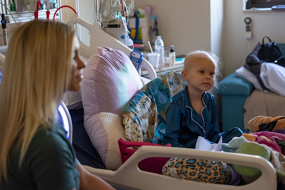 Mother and daughter listen to Marshall tell a story. The daughter sits in a hospital bed surrounded by stuffed animals and blankets.