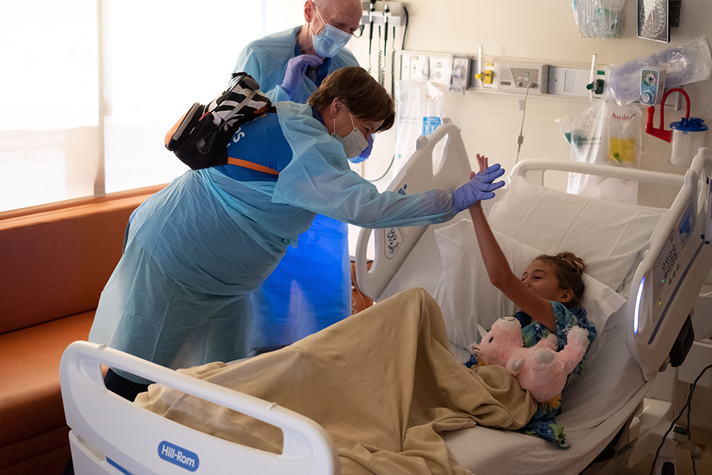 A Partner gives a high five to a girl in a hospital bed holding a pink stuffed unicorn